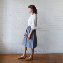 Load image into Gallery viewer, Woman wearing a white blouse and blue skirt standing against a white brick wall.
