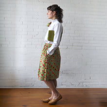 Load image into Gallery viewer, Person wearing a white shirt and patterned skirt against a white brick wall.
