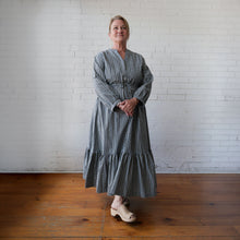 Load image into Gallery viewer, Woman wearing a long, checkered dress standing against a white brick wall.
