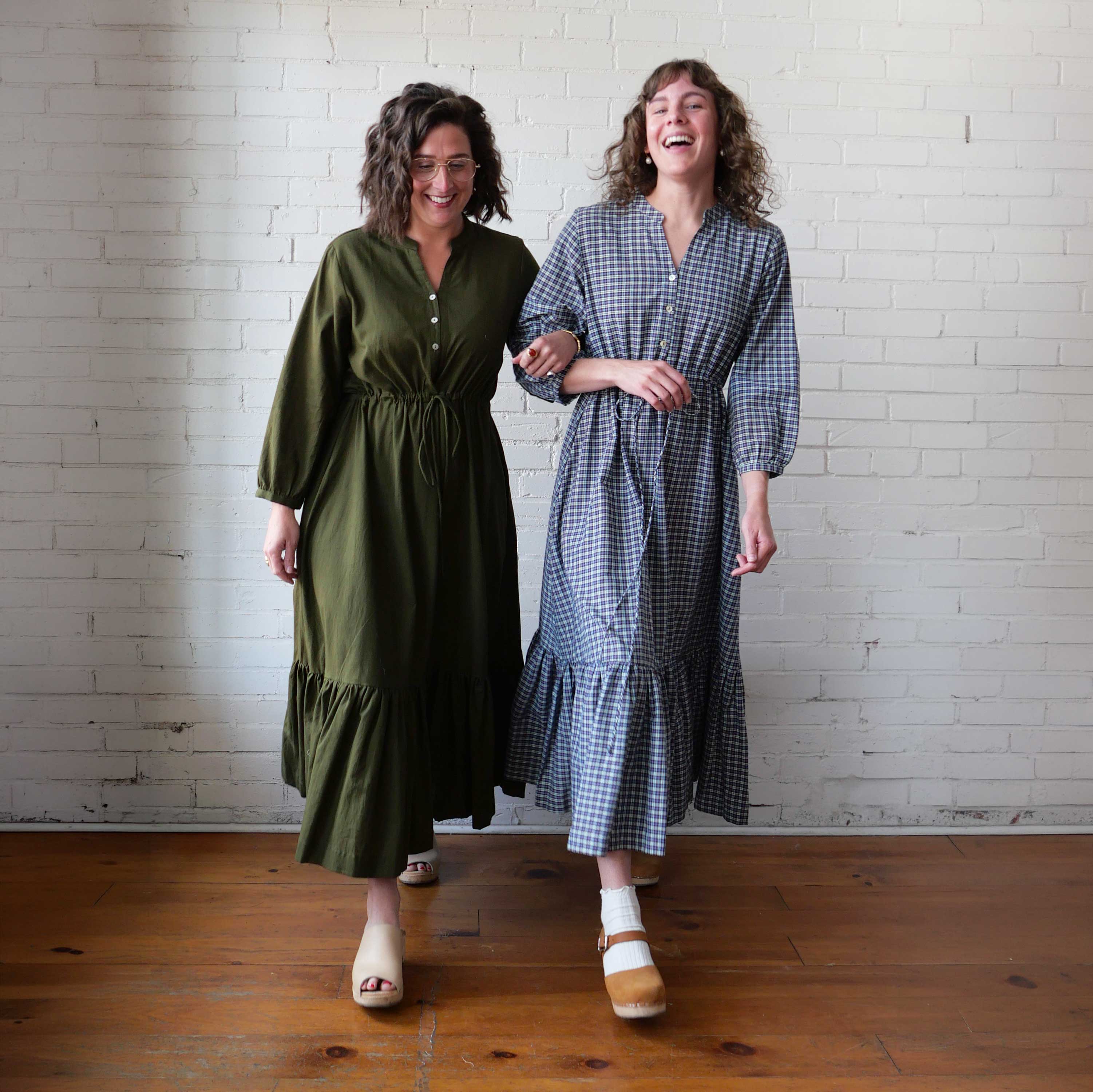 Two women standing side by side wearing long dresses against a white brick wall.