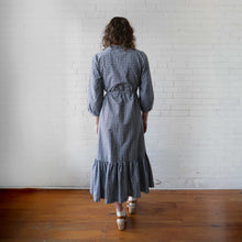Load image into Gallery viewer, Woman wearing a blue checkered dress standing against a white brick wall.
