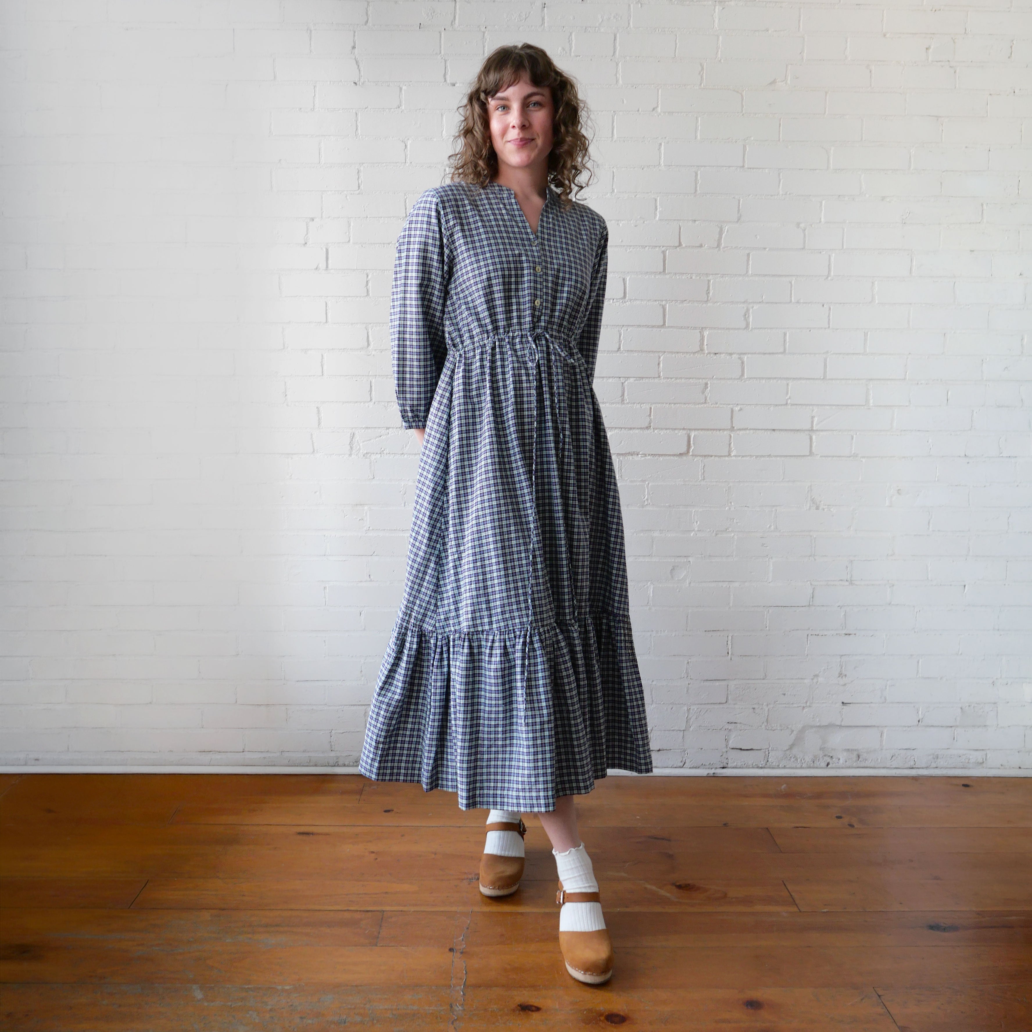 Woman wearing a blue checkered dress standing against a white brick wall.