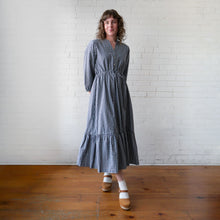 Load image into Gallery viewer, Woman wearing a blue checkered dress standing against a white brick wall.
