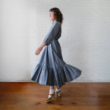 Load image into Gallery viewer, Woman wearing a blue and white checkered dress standing in front of a white brick wall.
