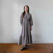 Load image into Gallery viewer, Woman wearing a checkered dress standing against a white brick wall.
