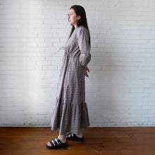 Load image into Gallery viewer, Woman wearing a checkered dress standing against a white brick wall.

