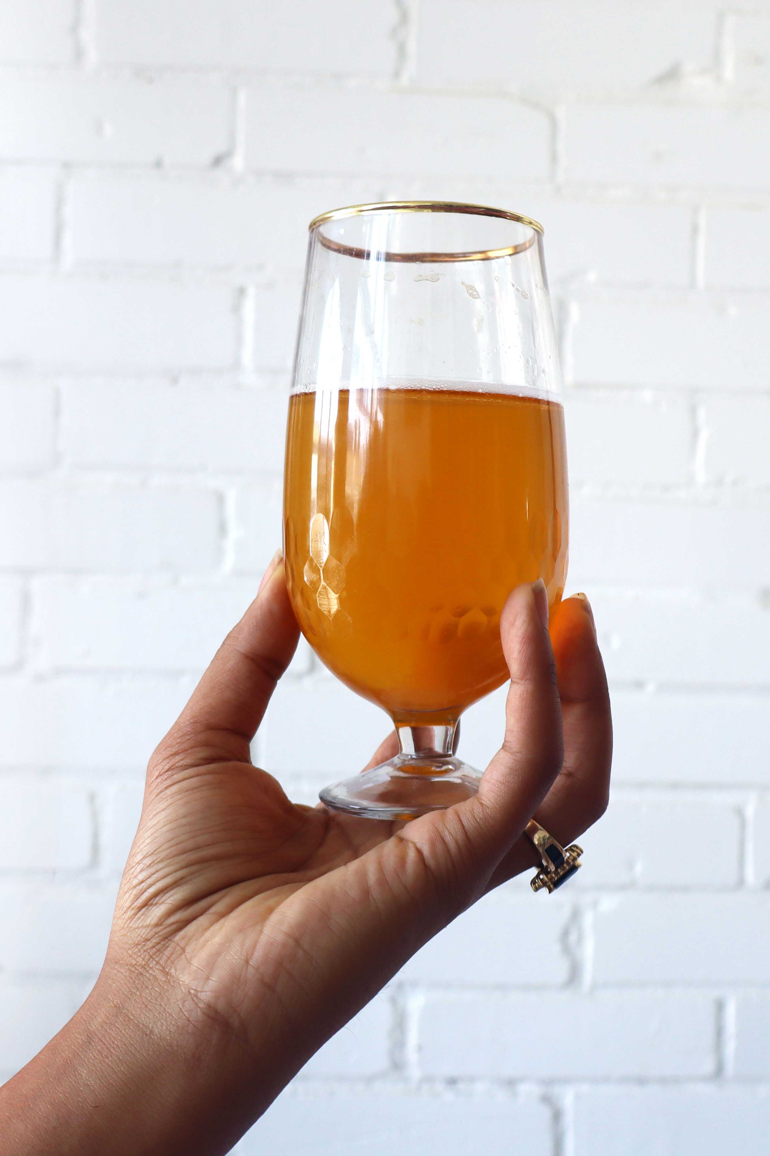 Hand holding a glass of orange beer against a white brick wall.