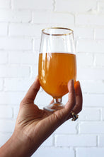 Load image into Gallery viewer, Hand holding a glass of orange beer against a white brick wall.
