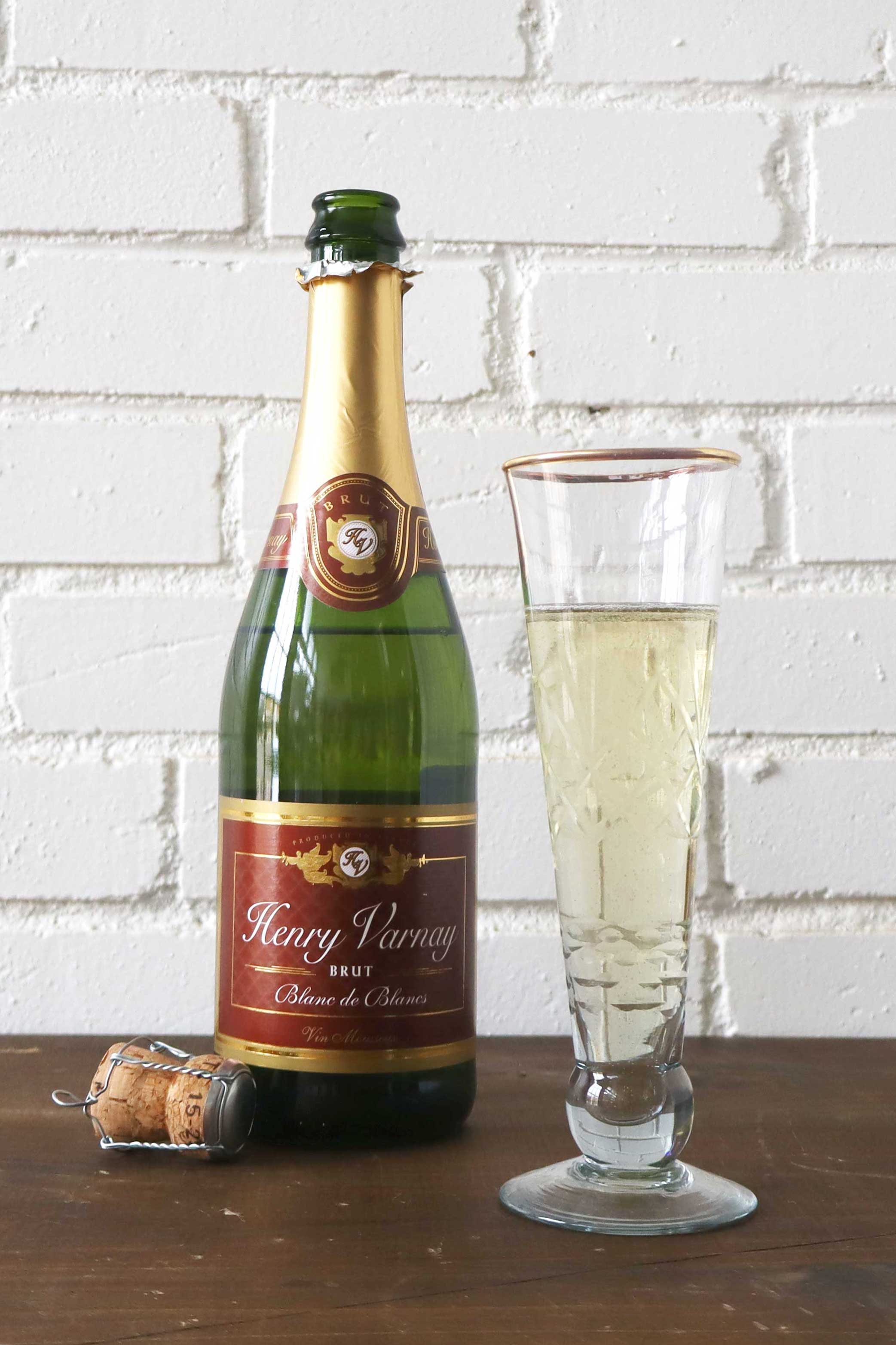Bottle of Henry Varnay wine with a glass of white wine on a wooden surface against a white brick wall.