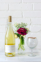 Load image into Gallery viewer, Wine bottle, vase with flowers, and wine glass on a white surface with a white brick wall background
